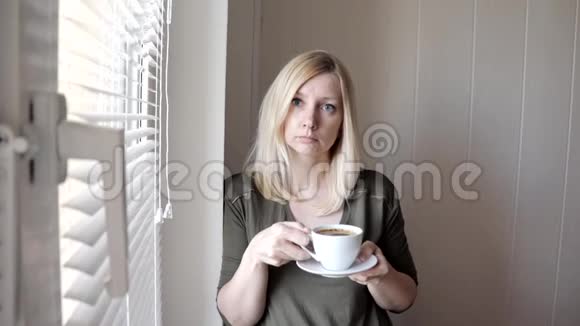 一个年轻的忧郁的美丽的金发女人早上站在窗户附近戴着百叶窗喝着咖啡视频的预览图