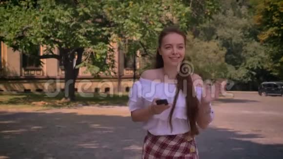 年轻女子一头姜发光着肩膀打电话走在大学大楼附近的公园里打招呼视频的预览图