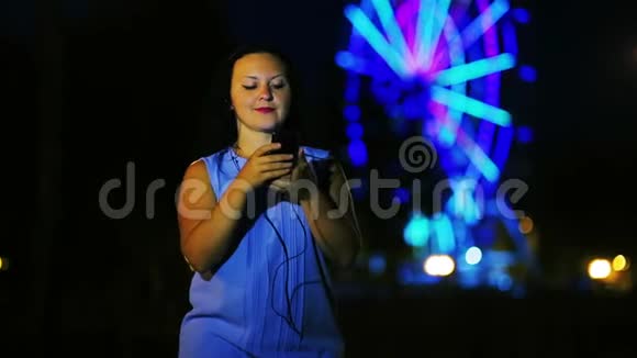一位戴着耳机的码头上的年轻女子在一个发光的摩天大楼的背景下在手机上看新闻视频的预览图