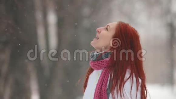 冬季公园站在人行道上的一个红头发的女人用嘴捕捉雪花视频的预览图