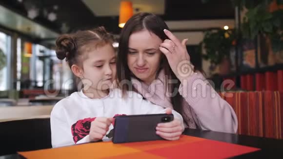 漂亮的白种人母亲和她的女儿在咖啡馆里使用智能手机一个先进的小女孩展示并解释视频的预览图