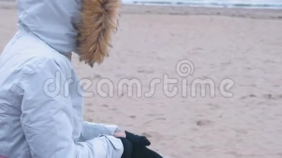穿着白色羽绒服的女人坐在海滩上的长凳上戴上手套海冬冻女人视频的预览图