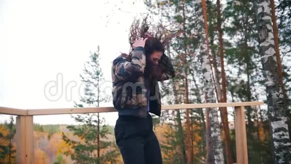 微笑的年轻女子在平台上跳舞和跳跃会飞的头发背景上的森林视频的预览图