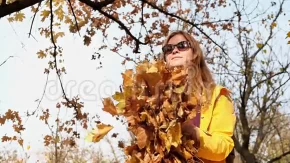 穿着黄色雨衣的年轻金发女子在公园里快乐地扔秋叶视频的预览图