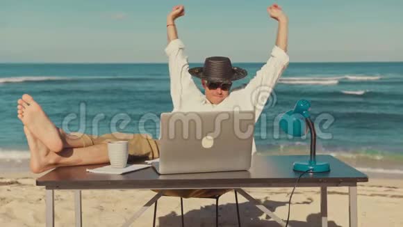 一个年轻人拿着笔记本电脑坐在海滩上休息着把腿放在桌子上视频的预览图