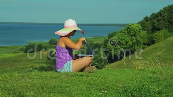 戴着草帽的女人坐在俯瞰湖面的山上用笔记本电脑工作视频的预览图