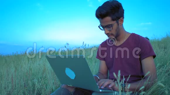 戴眼镜的印度男子在一天的日落时分坐在草地上的笔记本电脑后面工作视频的预览图
