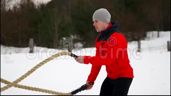 年轻的肌肉男穿着红色夹克训练着战斗绳冬天和雪慢动作户外健身运动视频的预览图