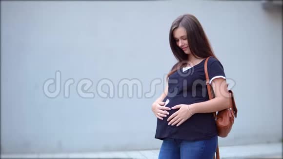 一个穿着T恤和背包的漂亮怀孕女孩在灰色的背景上抚摸着她的肚子视频的预览图