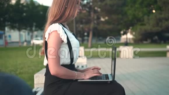 白人商务女性穿着白色衬衫和眼镜坐在街上的长凳上在笔记本电脑后面工作视频的预览图