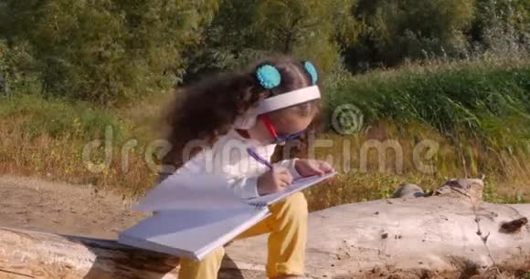 快乐的小女孩坐在河边听着耳机上的音乐在平板电脑上画画视频的预览图