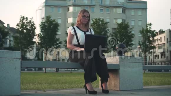 穿着白色衬衫和眼镜的白人年轻商务女性坐在街上的长凳上在笔记本电脑后面工作视频的预览图