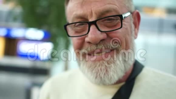 一个戴着眼镜和灰色胡须的成熟男人的亲密肖像退休人员在机场微笑购物视频的预览图