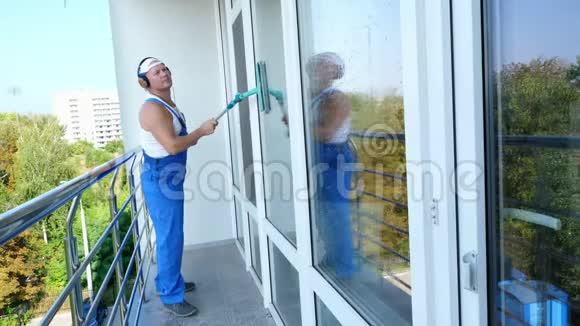 白人帅哥清洁工穿着蓝色工作服带着耳机清洗窗户视频的预览图