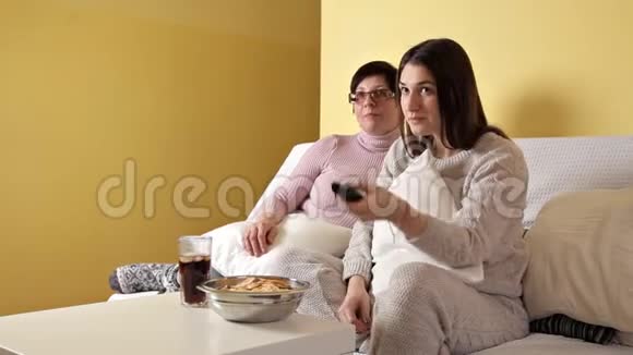 年轻女孩和她妈妈在家沙发上看电视吃薯片喝可乐晚上好房子舒适舒适视频的预览图