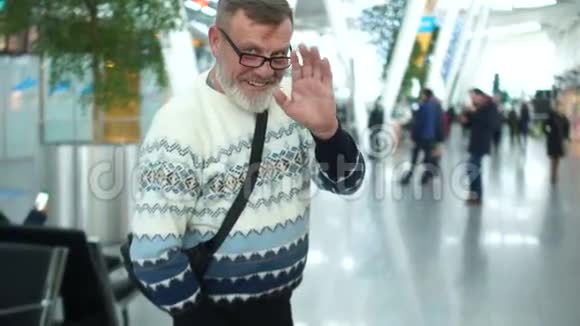 在机场或火车站穿着针织毛衣和眼镜胡子灰白的成熟男人养老金领取者挥手视频的预览图