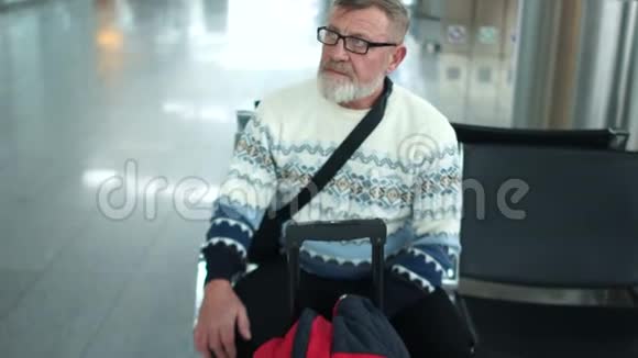 成熟迷人的男人穿着白色毛衣和眼镜坐在机场休息室他们宣布了他的航班他站起来视频的预览图