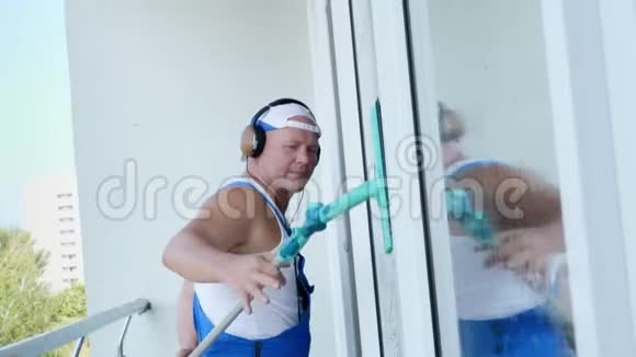 白种人有魅力的男人清洁服务工人穿着带耳机的蓝色工作服用特殊的方式清洗窗户视频的预览图