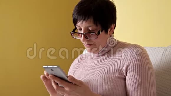 一个戴眼镜的女人在家里抱着一个白色枕头坐在沙发上在她全新的智能手机上写着信息视频的预览图