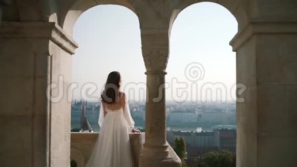 一位非常漂亮的年轻女子或新娘穿着白色连衣裙呆在老堡的阳台上看着城市视频的预览图