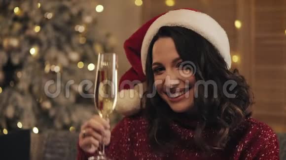 迷人的女人戴着圣诞帽的香槟看着圣诞树背景上的相机她养大了视频的预览图