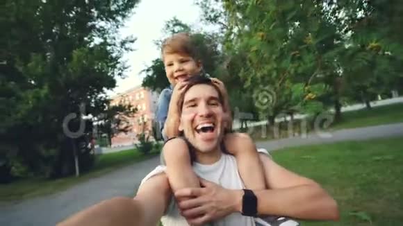 快乐的父亲带着他微笑的儿子在公园散步时在智能手机摄像头上自拍视频的预览图