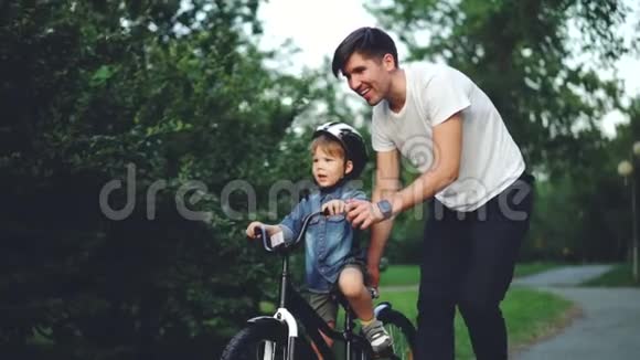 快乐的年轻人爱父亲的慢动作在夏天的绿色公园里教孩子骑自行车小男孩在笑视频的预览图