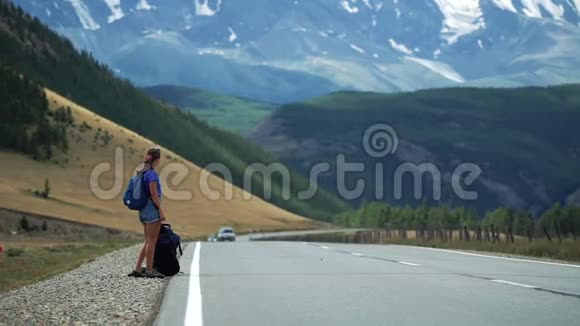 旅行者女孩带着背包和短裤在山路上搭便车那里有雪山视频的预览图