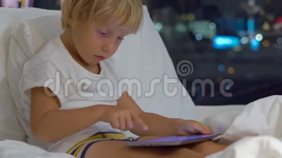 一个晚上坐在床上的小男孩近距离拍摄了一台平板电脑电脑上有摩天大楼的轮廓视频的预览图
