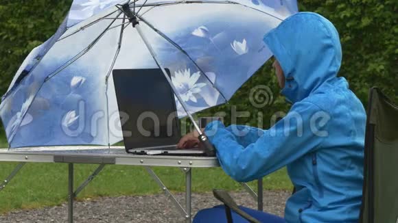 穿防水衣服的商务女士在雨中的野餐桌上工作笔记本电脑视频的预览图