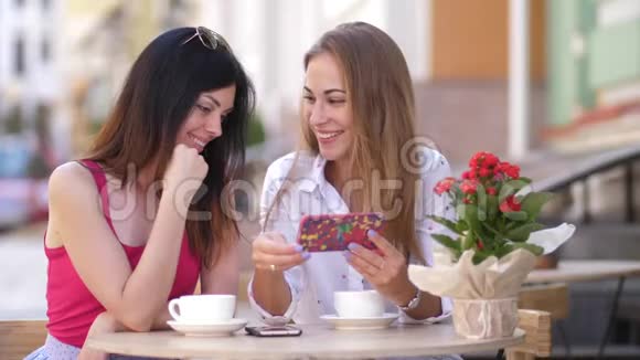 两个漂亮的年轻女人坐在咖啡馆的桌子旁看着电话4k慢动作视频的预览图