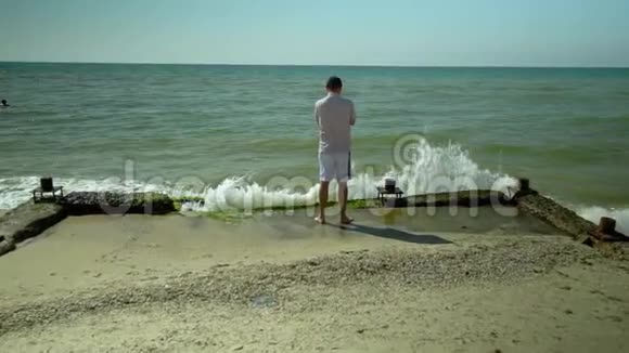 一个年轻人站在海滩上看着海浪海浪拍打着海岸慢动作视频的预览图