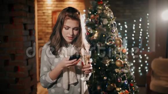 一个手里拿着一杯香槟的漂亮女孩正笑着看着圣诞节手机上的照片视频的预览图