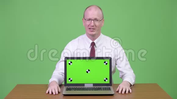 成熟的秃头商人一边说话一边拿笔记本电脑对着木桌视频的预览图