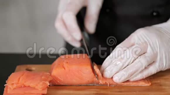 一位年轻的男寿司厨师用一把刀在桌子上切下一个熏鲑鱼锉刀双手戴手套特写视频的预览图
