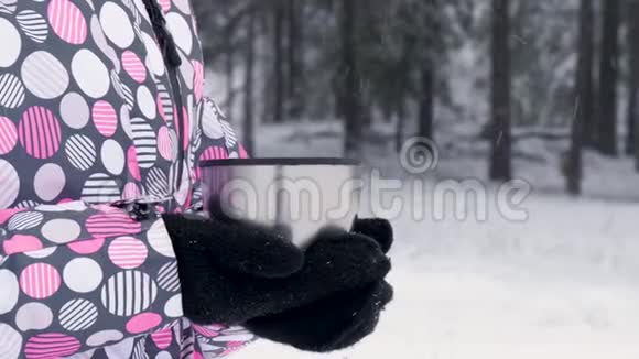 在一个下雪的日子里戴手套的女性手在森林的背景下捧着一杯茶或咖啡视频的预览图