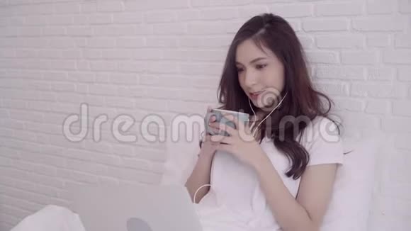 一幅美丽迷人的亚洲女人在床上躺着时用电脑或笔记本电脑拿着一杯温暖的咖啡或茶的肖像视频的预览图