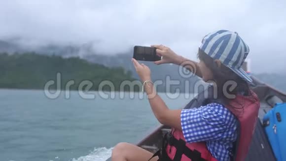 年轻女子乘坐快速移动的旅游木船携带手提箱在多云间阴天用手机拍照视频的预览图