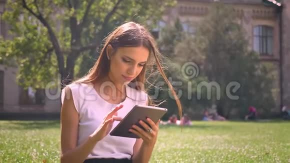 女士白天坐在公园的草地上敲击平板电脑放松概念交流概念视频的预览图