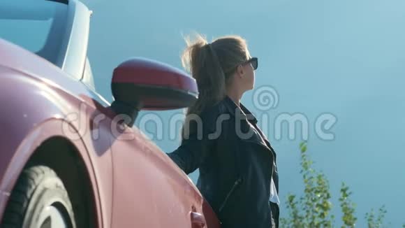 穿着黑色皮夹克的年轻女子在天空背景下摆着红色敞篷车视频的预览图