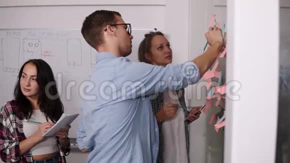 由三个年轻人组成的团队戴眼镜的年轻人把彩色的纸粘在一起并在上面做一个笔记同事视频的预览图