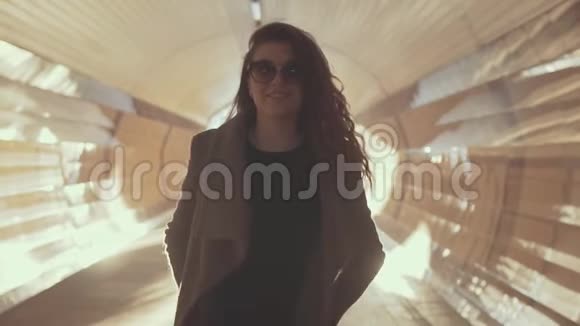 戴着太阳镜的迷人少女在夕阳下穿着外套穿过阳光明媚的隧道视频的预览图
