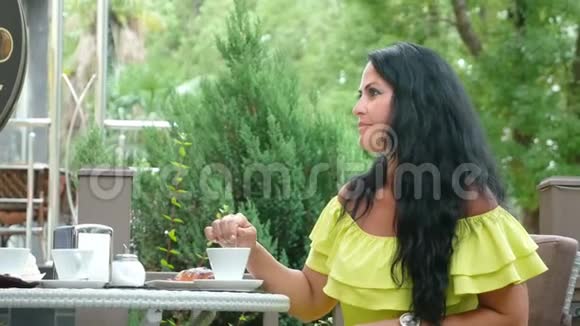 年轻漂亮的黑发女人坐在户外咖啡馆里喝着咖啡吃着羊角面包视频的预览图