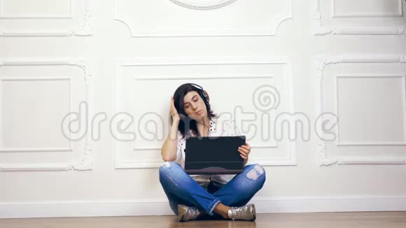 年轻女性女孩深色头发穿着白色衬衫和牛仔裤坐着听音乐使用耳机和笔记本电脑视频的预览图