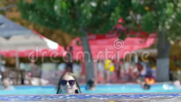 在蓝色的夏日游泳池里微笑的女孩戴上太阳镜的肖像视频的预览图