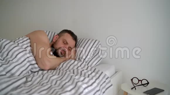 大胡子的男人在毯子下面的床上醒来微笑着戴上眼镜拿着他的智能肾视频的预览图