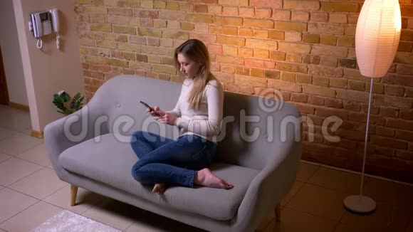 一位年轻迷人的白种人深褐色皮肤的女性在一个舒适的室内沙发上发着手机短信她的特写照片很吸引人视频的预览图