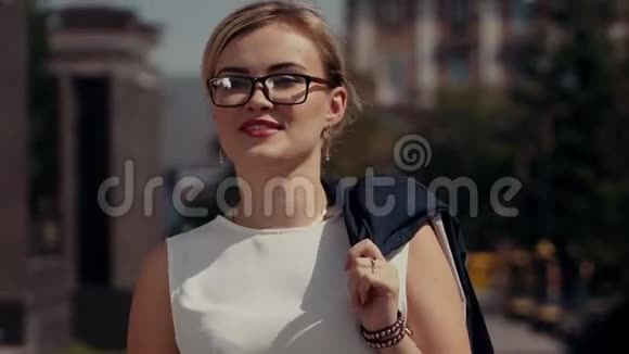 一个戴着眼镜和金发的年轻女孩正沿着城市阳光明媚的街道走着在她后面的建筑后面视频的预览图