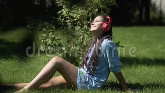 在阳光明媚的天气里一个留着长发的女孩坐在公园的草地上用智能手机听耳机里的音乐视频的预览图