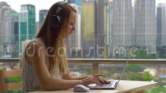 年轻女性自由职业者在阳台上的笔记本电脑上工作背景是一个充满摩天大楼的市中心远程视频的预览图